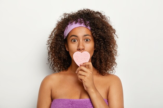 Photo of attractive young Afro American woman wears purple headband and towel around body, covers mouth with cosmetic heart shaped sponge, wipes face