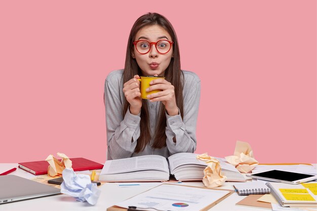 Photo of attractive young female holds mug with hot beverage, wears big spectacles, dressed in stylish shirt