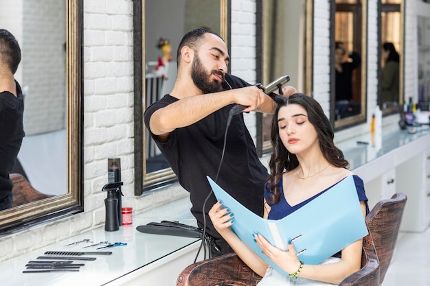 Photo at the barbershop young Barber trying to form her clients hair and client looking at the catalog High quality photo