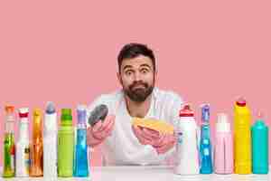 Free photo photo of bearded atrractive busy husband holds two sponges, raises eyebrows with surprisement, wears rubber protective gloves