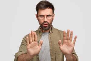 Photo of bearded young male shows stop gesture, has displeased facial expression, denies something, talks about forbidden things, wears fashionable shirt, isolated over white wall