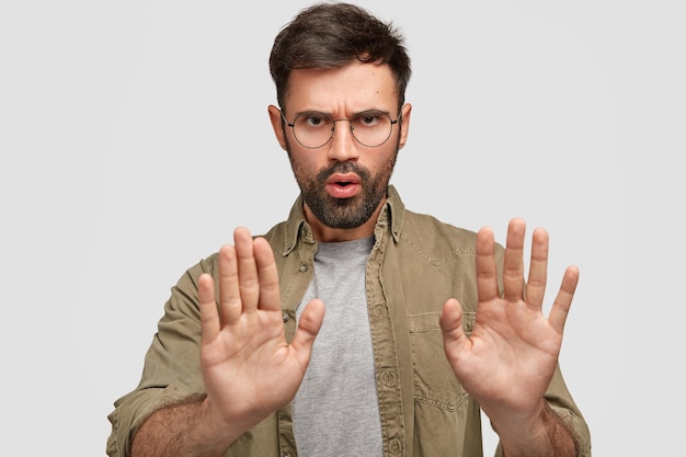 Фотография молодого бородатого мужчины показывает стоп-жест, недовольное выражение лица, что-то отрицает, говорит о запретных вещах, носит модную рубашку, изолирована на белой стене