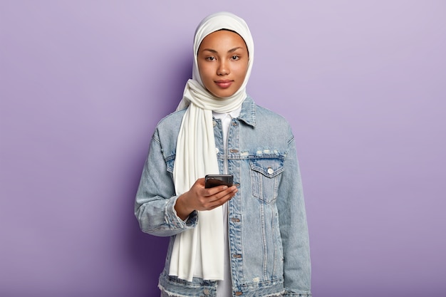 Foto gratuita foto di una bella donna araba con la pelle scura e sana, un messaggio di testo sul cellulare moderno, legge i commenti sotto il post, indossa hijab bianco e modelli di cappotto di jeans sul muro viola. religione musulmana