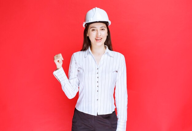 赤い壁に立っている安全帽子をかぶった美しいビジネス女性の写真。