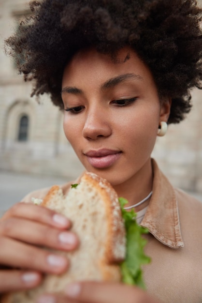 Photo of beautiful curly haired woman eats sandwich outdoors likes fast food feels very hungry poses at street dressed in casual clothes. Lunch break outside. People and unhealthy nutrition concept