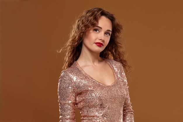 Photo of beautiful seductive young caucasian woman in golden cocktail dress looking into the camera on brown background