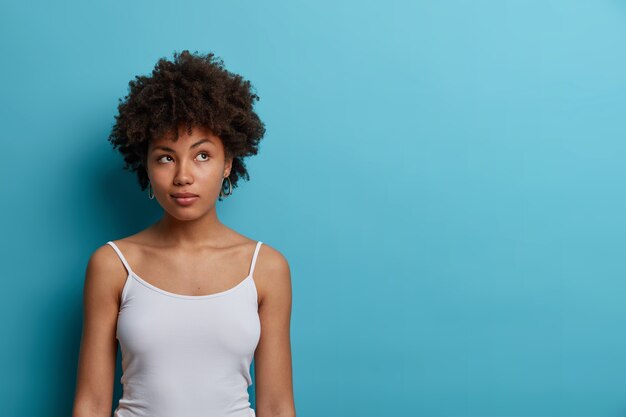 Photo of beautiful thoughtful woman ponders about something, looks pensively aside, has some ideas in mind, dressed casually, isolated on blue wall, blank space for your advertisement.