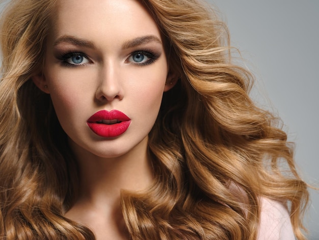 Photo of a beautiful young blond woman with sexy red lips. Closeup attractive sensual face of  girl with long curly hair. Smoky eye makeup