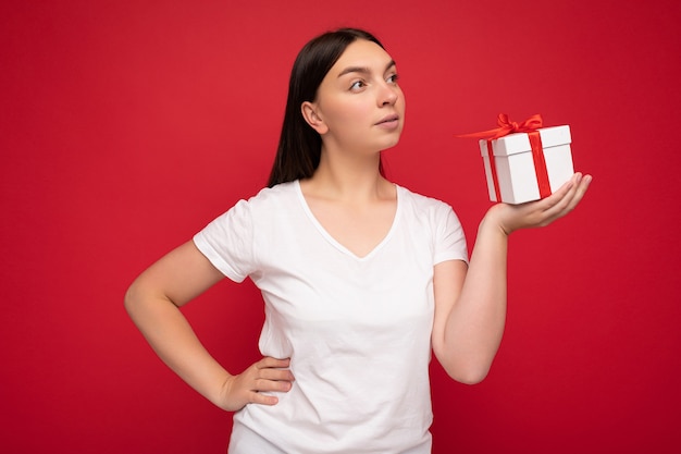 Photo of beautiful young brunette woman isolated over red background wall wearing white casual t-shirt for mockup holding white gift box with red ribbon and looking to the side Premium Photo