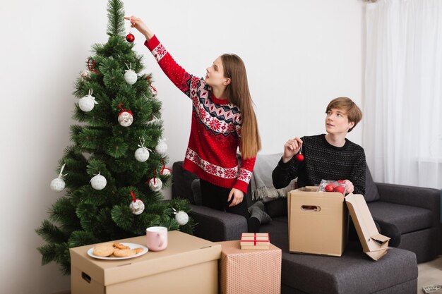 Photo of beautiful young couple sitting on sofa at home and dreamily decorating Christmas tree together