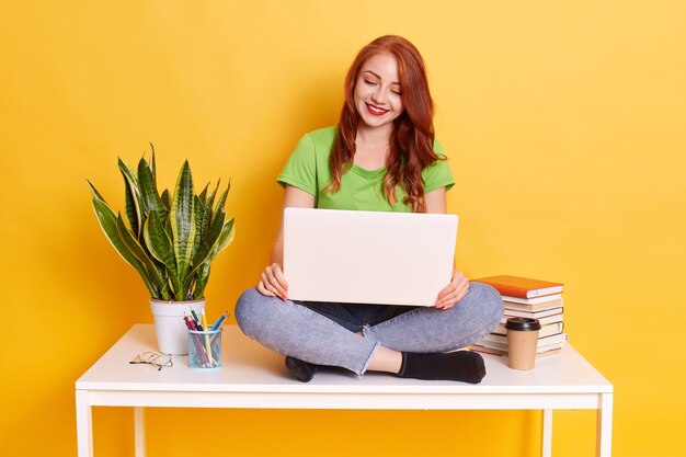 캐주얼 옷을 입고 무릎에 무릎 위로 흰색 테이블에 앉아, 온라인 작업 또는 학습 거리에 앉아 아름 다운 젊은 빨간 머리 대학생의 사진.