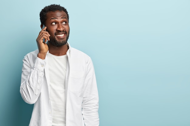 Photo of black ethnic hipster guy has phone conversation, holds mobile phone near ear, tells news to friend, focused away, wears white shirt. Successful businessman makes appointment via cellular