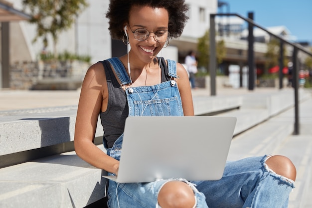 Photo of black glad woman views web pages, keyboards on laptop computer feedback or comments, listens online broadcasting in earphones, wears ragged dungarees, does remote work, models outside