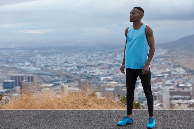 Photo of black sportsman wears blues sneakers, vest and leggings, models against altitude above horizon, big city and mountains, free space for your information. Panoramic view