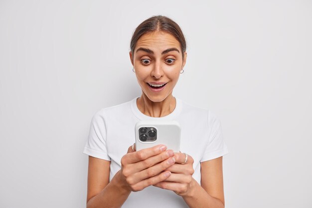 Photo of brunette woman reads received message with bated breath stares at mobile phone screen gets amazing offer feels thrilled has eyes popped out wears casual t shirt isolated on white wall