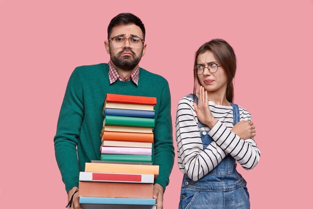 Photo of busy bearded male student carries many books, displeased female shows disagreement gesture