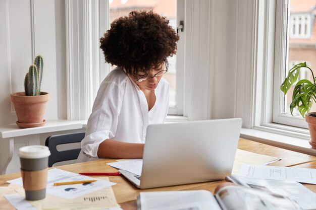 Photo of busy dark skinned manager talks with customer via cell phone, writes down some information in papers, sits at desktop and notes text, laptop computer