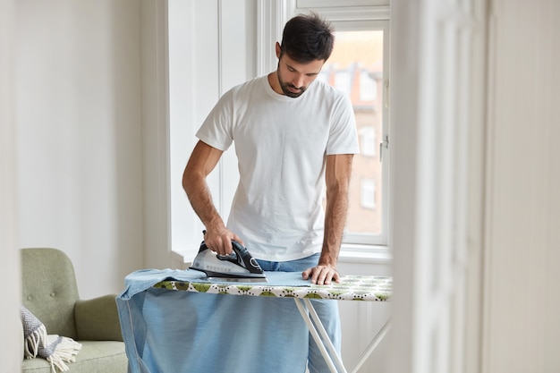 Photo of busy unshaven man irons shirt on ironing board, prepares for formal meeting on business conference