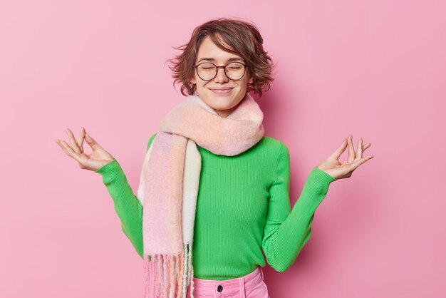 Photo of calm concentrated woman meditates with closed eyes and zen fingers tries to find inner balance enjoys meditation wears green jumper warm scarf around neck isolated on pink wall Yoga concept