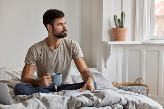 Photo of calm handsome unshaven man enjoys reading bestseller, holds mug with coffee or tea, sits crossed legs at bed, ponders on life situation, looks pensively aside. People and hobby concept