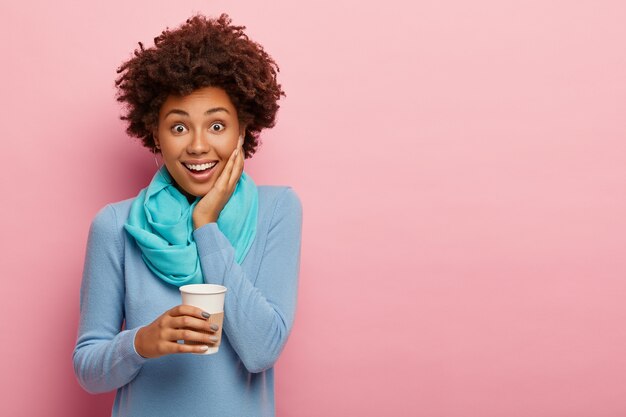 Photo of carefree curly woman holds disposable cup of coffee, enjoys drinking aromatic beverage, wears blue casual clothes, has free time after work, poses over rosy wall, copy space for your advert