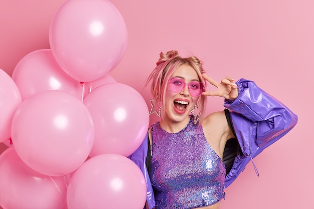 Photo of carefree joyful woman has fun on birthday party wears trendy sunglasses and purple jacket exclaims with happiness makes peace gesture holds bunch of inflated balloons poses over pink wall