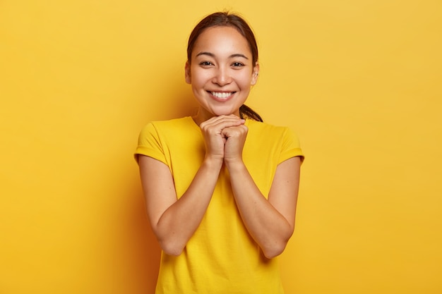 На фото харизматичная азиатка держит руки вместе около подбородка, нежно улыбается, у нее милое выражение, темные волосы зачесаны в хвост, носит ярко-желтую футболку, развлекается в отличной компании.