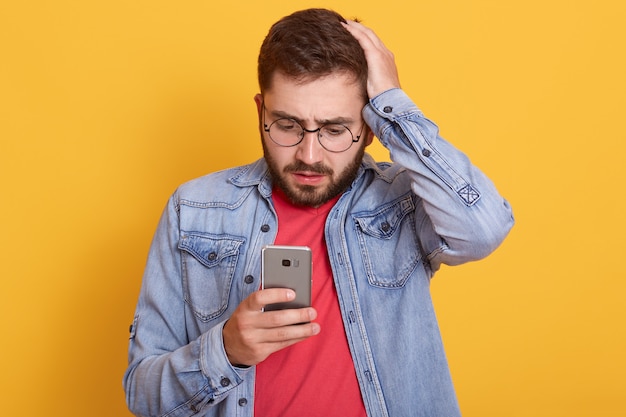 Photo of charismatic shocked young bearded man holding his smartphone, looking at device screen attentively, putting on hand on head