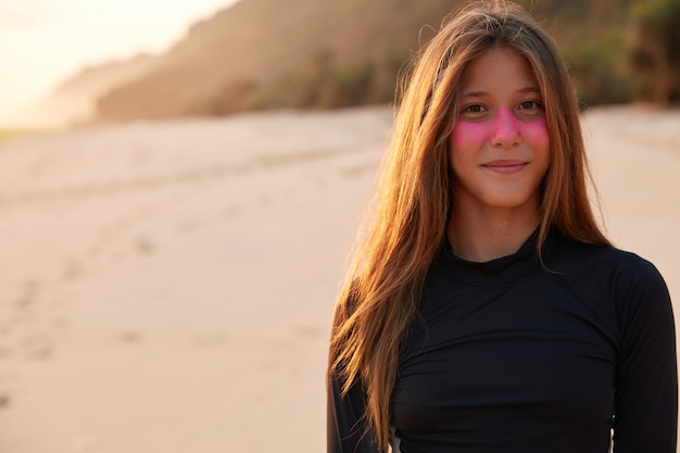 Photo of charming woman surfer protects face with zinc mask, has outdoor stroll against sandy beach