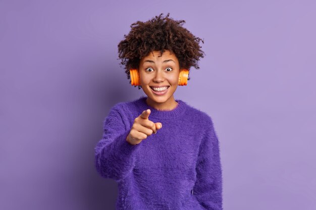 쾌활한 아프리카 계 미국인 여자의 사진은 행복하게 미소 짓고 카메라를 직접 가리키며 귀에 헤드폰을 착용하고 좋아하는 음악을 듣는 것을 선택하면 매우 기쁩니다.