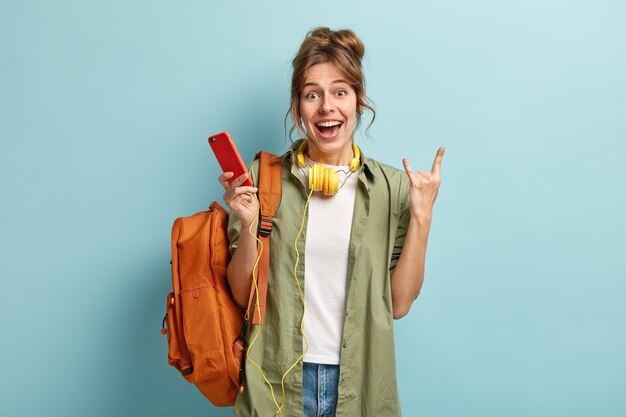 Photo of cheerful Caucasian woman makes horn gesture, holds smart phone, listens audio book