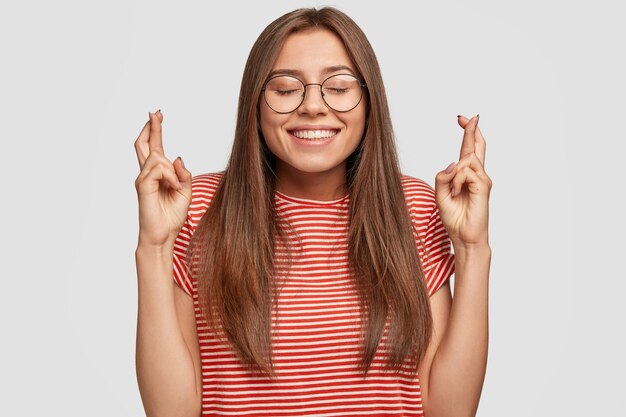 Photo of cheerful dark haired woman believes in good fortune, keeps fingers crossed