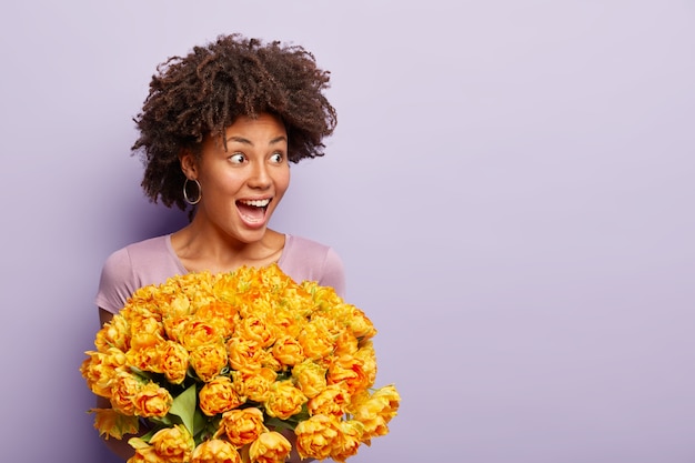 Фотография веселой темнокожей женщины с свежими волосами, держит оранжевые тюльпаны, носит повседневную футболку, выражает счастье, позирует над фиолетовой стеной, свободное место для вашей рекламы. Подруга получает цветы
