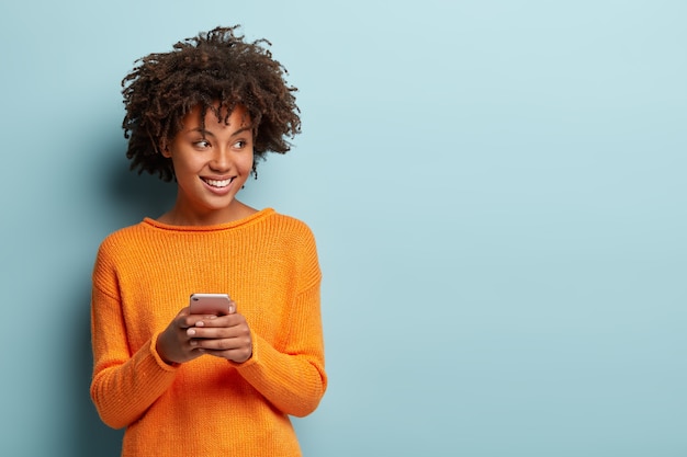 Photo of cheerful delighted African American woman types sms on modern cell phone device, enjoys good internet connection