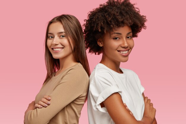 明るく多様でフレンドリーな女性の写真は、お互いに立ち向かい、手を組んで、コラボレーションの結果に満足し、カジュアルな服を着て、ピンクの壁に隔離されています。横ショット