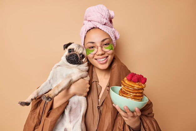 Photo of cheerful dog owne poses with favorite pet and pancakes with syrup and raspberries applies green patches under eyes wears nightwear wrapped in blanket isolated over beige background