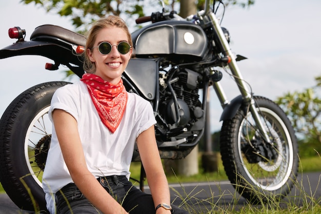 Photo of cheerful female biker sits near black motorcycle in open air, wears stylish clothes, travels in unknown countryside place against wonderful scene. Outdoor lifestyle concept.