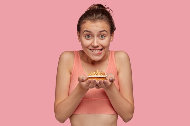 Photo of cheerful fitness lady feels temptation while holds delicious sweet cake, wears casual top, bites lips, doesnt eat dessert as wants to be in good shape, isolated over pink wall