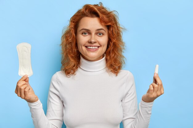 Photo of cheerful good looking female holds means of protection during menstruation, holds cotton clean sanitary napkin and tampon, controls menstrual cycle, wears white jumper. Personal care