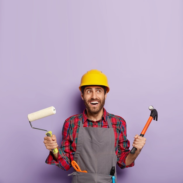 Photo of cheerful male builder or decorator holds hammer and paint roller, ready for painting walls and repairing, has glad expression, looks above.