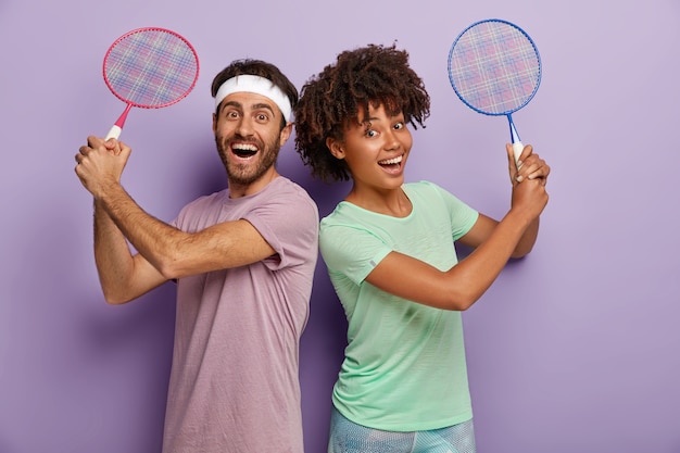 Free photo photo of cheerful mixed race woman and man stand back to each other, hold tennis racket, enjoy playing, laugh positively, dressed in t shirts, being active and satisfied