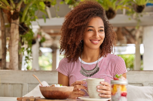 Фотография веселой расслабленной черной девушки с кудрявыми волосами, держащей кружку кофе, увлекающейся времяпрепровождением, посещающей экзотический кафетерий, летних отпусков за границей, смотрит в сторону