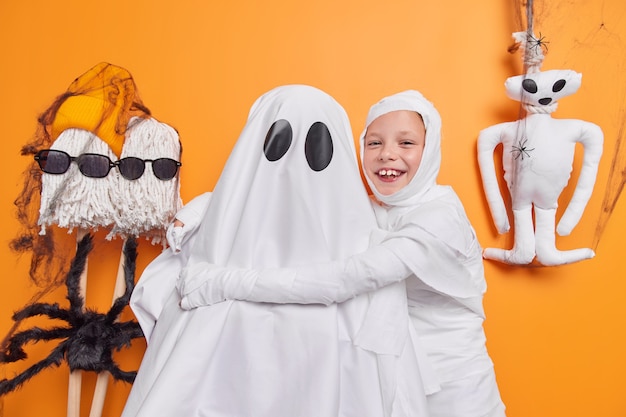Free photo photo of cheerful small child embraces ghost has fun foolishes around poses on orange