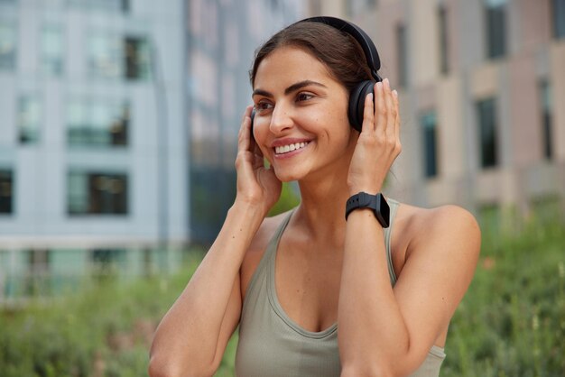 Photo of cheerful sporty woman keeps hands on headphones strolls on urban street looks happily into distance uses smartwatch for fitness tracking has happy expression listens favorite music.