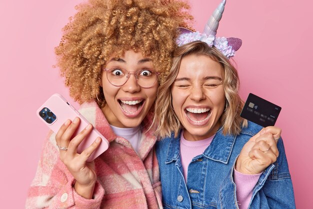 Photo of cheerful two female friends glad to get salary spend money on online shopping holds mobile phone and banking card dressed in stylish jackets make purchases in internet isolated on pink wall