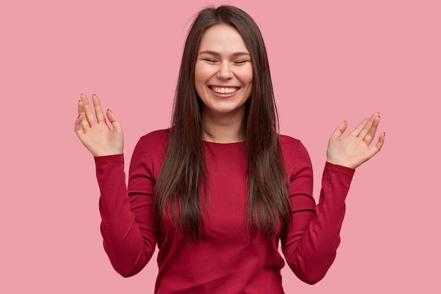 Photo of cheerful woman with toothy smile, closes eyes from pleasure, raises palms, expresses positive feelings, stands against pink background
