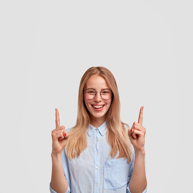 Photo of cheerful young cute female with gentle smile, indicates upwards with both index fingers, shows something above head, wears elegant-shirt and spectacles, isolated over white wall