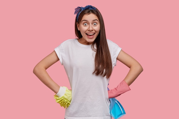 Photo of cheerful young housemaid keeps hands on waist, wears casual white t shirt, headband, protective gloves