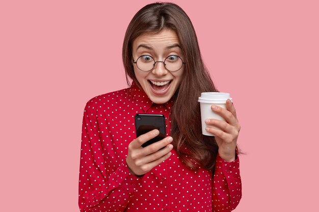 Photo of cheerful young woman checks social networks, plays games on phone, browses online apps, drinks takeaway coffee, dressed in red clothes