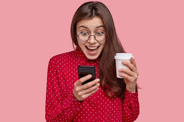 쾌활한 젊은 여성의 사진은 소셜 네트워크를 확인하고, 전화로 게임을하고, 온라인 앱을 탐색하고, 빨간 옷을 입은 테이크 아웃 커피를 마신다.